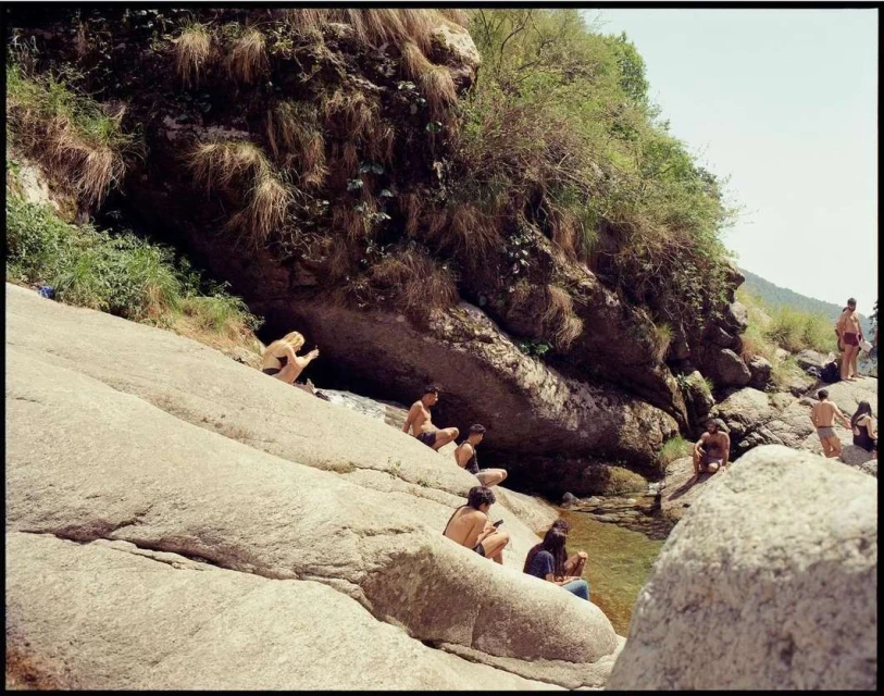 People are relaxing on the rocks near a mountain stream. The water is running over the rocks, and there is vegetation on the surrounding rocks.