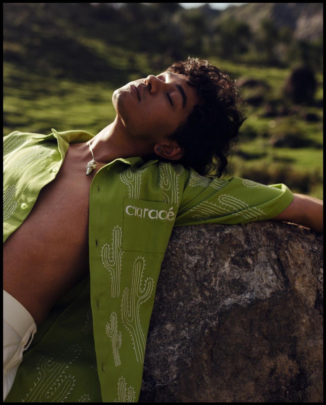 Young man with curly hair relaxes with eyes closed on a rock wearing a light green cactus-print shirt and tan pants outside.