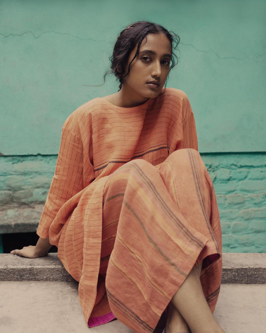 A South Asian woman is sitting on a concrete surface, wearing a striped peach dress, with a turquoise wall in the background.