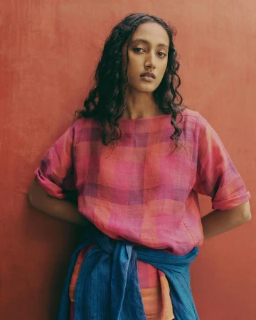 Portrait of a dark-haired woman wearing a pink plaid shirt and blue sash against an orange backdrop in a studio setting.