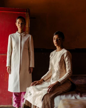 Two women in elegant cream-colored dresses pose indoors against a red door and brown wall in a fashion editorial shot with natural light.