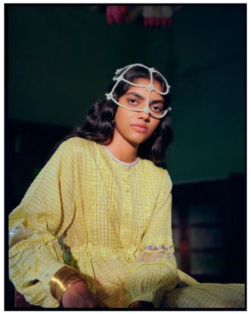 Portrait of a young woman with dark hair wearing a yellow dress and elaborate beaded headpiece, posed against a dark green background.