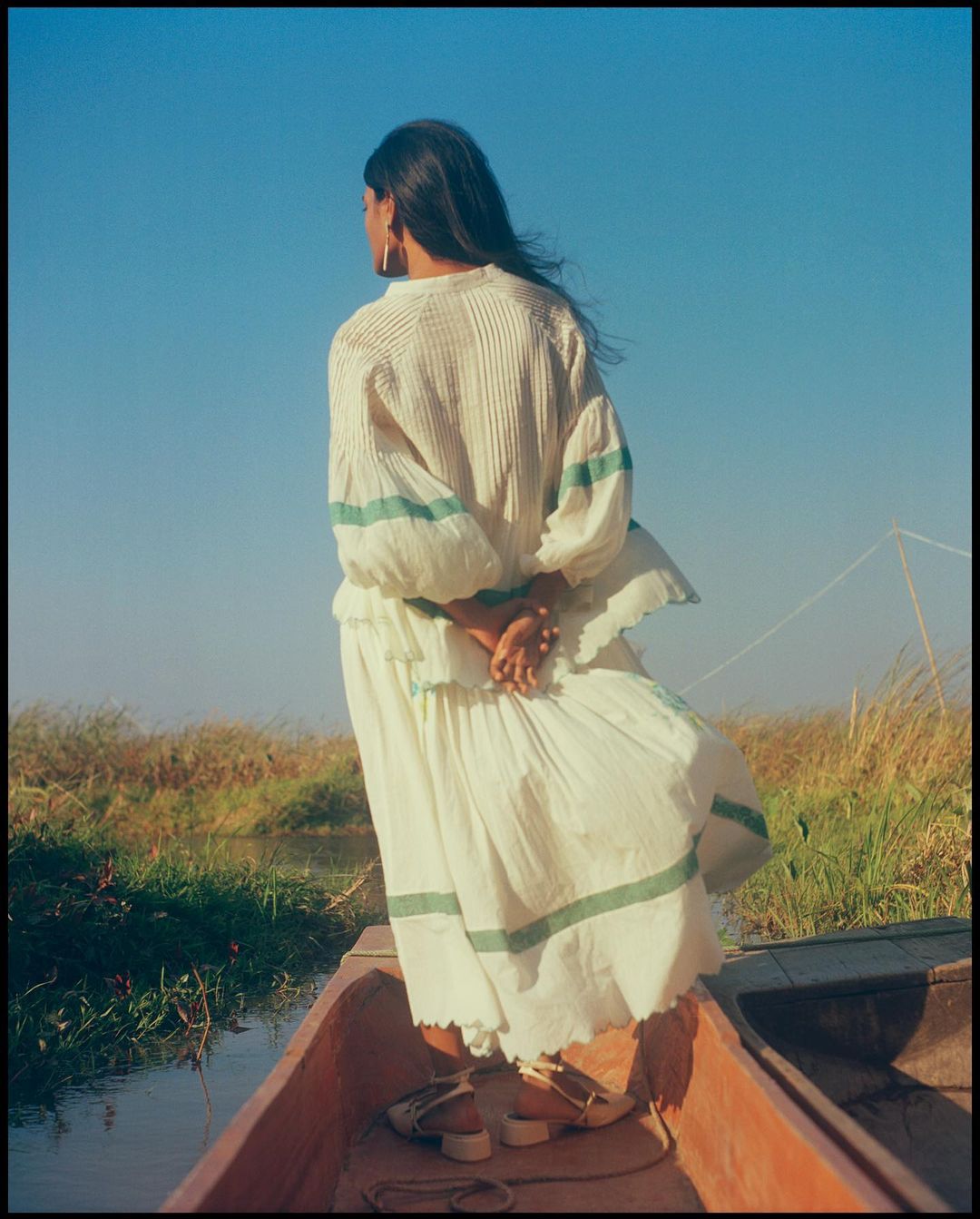 Woman in flowing white dress stands in wooden boat against blue sky with marshy landscape. Calm waters, serene ambiance.