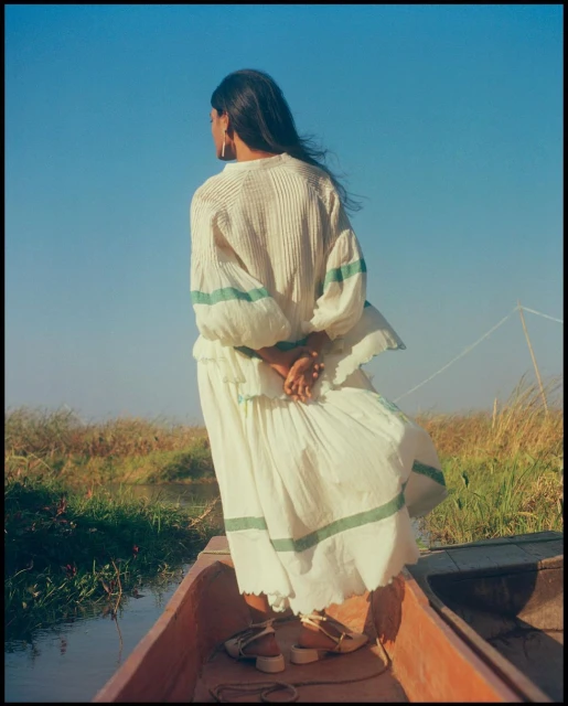 Woman in flowing white dress stands in wooden boat against blue sky with marshy landscape. Calm waters, serene ambiance.