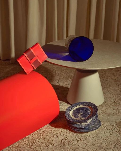 Still life photograph of a red clutch handbag on an orange cylinder, with a blue disc, plates and a white table, against neutral curtains.