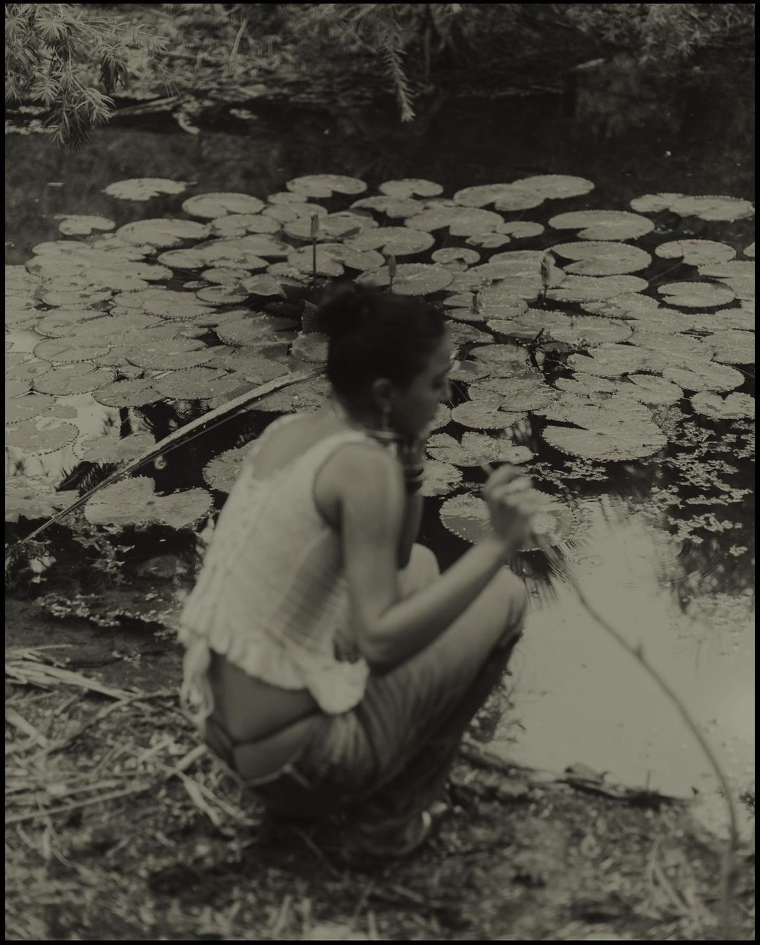 Woman crouches by a pond with lily pads, holding a long object, in a vintage-style black and white outdoor photograph. 