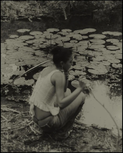 Woman crouches by a pond with lily pads, holding a long object, in a vintage-style black and white outdoor photograph. 