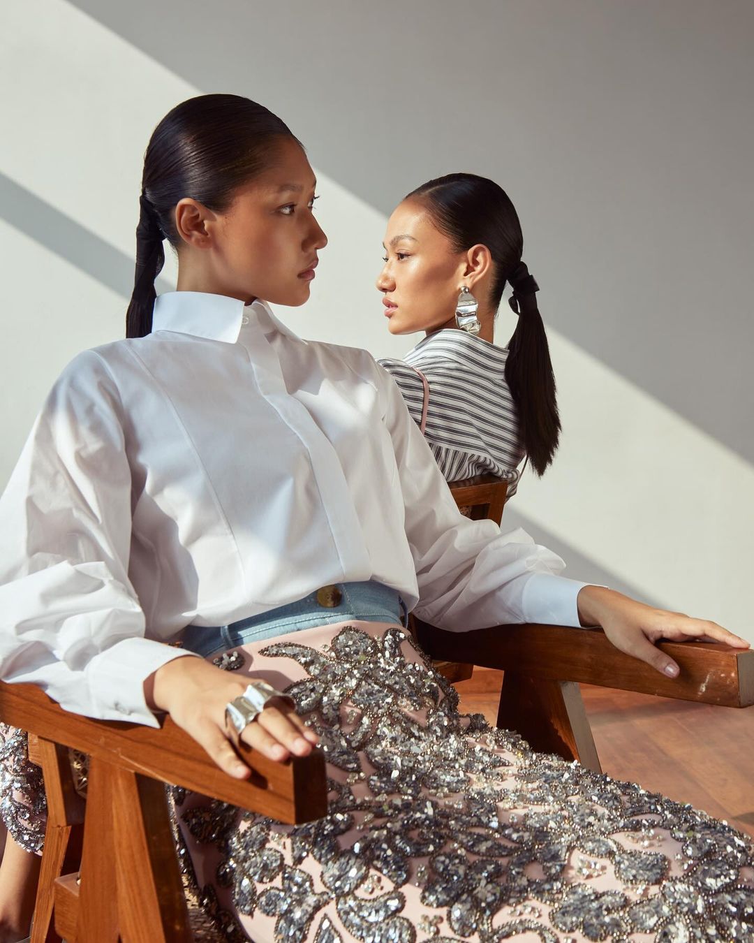Two Asian women in a photo studio wearing elegant white shirts, dark ponytails, and silver sequined skirts, sitting on wooden chairs.