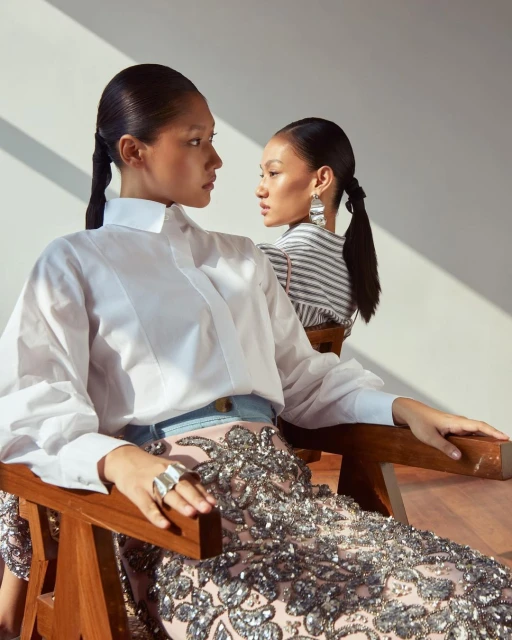 Two Asian women in a photo studio wearing elegant white shirts, dark ponytails, and silver sequined skirts, sitting on wooden chairs.