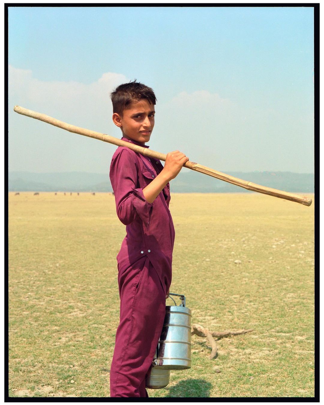 A young boy in maroon clothes carrying a long wooden stick and stacked lunch containers in a field with a blue sky background.