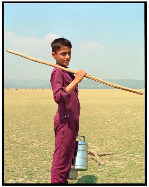 A young boy in maroon clothes carrying a long wooden stick and stacked lunch containers in a field with a blue sky background.