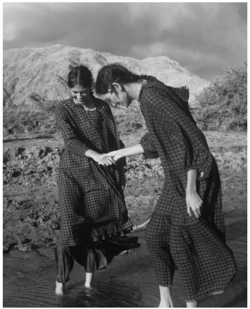 Two women in vintage dresses stand in shallow water, holding hands against a backdrop of mountains and cloudy skies in black and white.