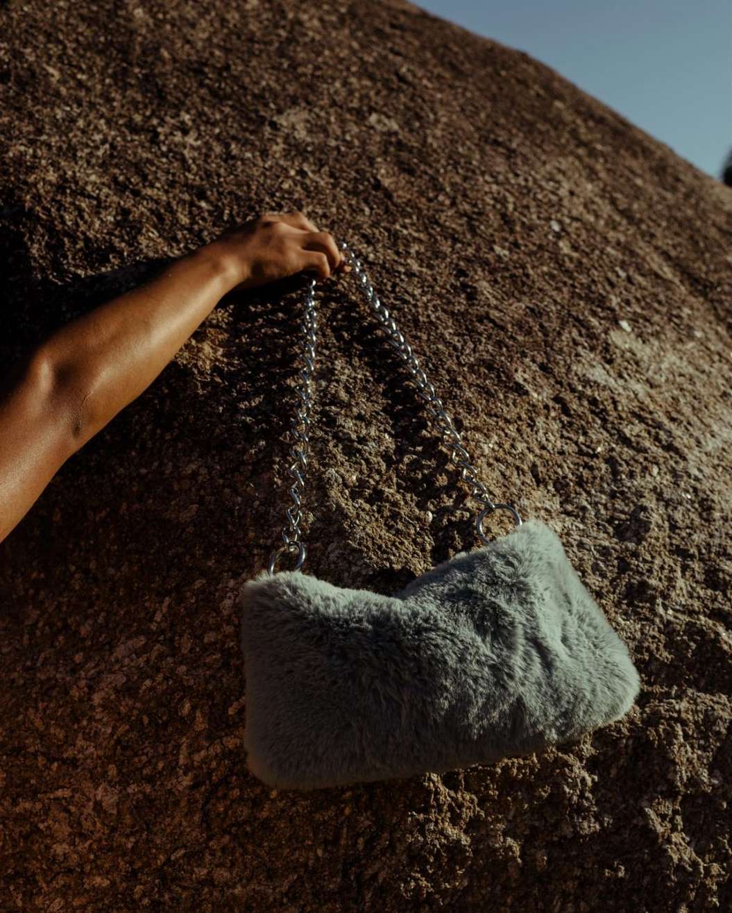 An arm holds a fur handbag with a silver chain strap over a large brown rock formation outdoors, lit by sunlight.