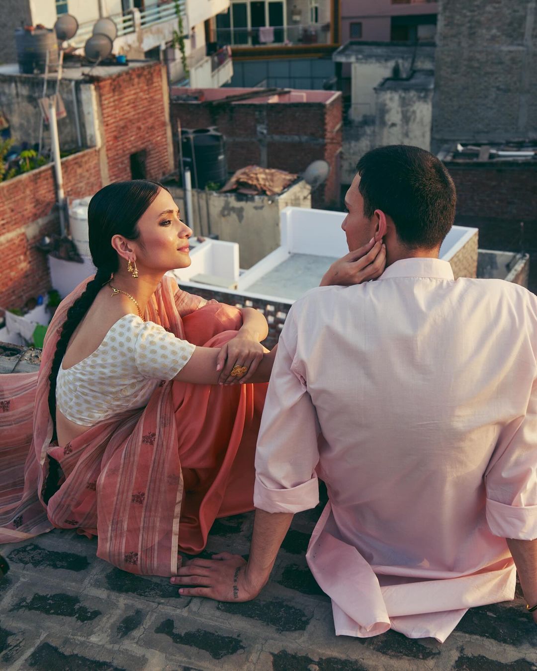 Romantic couple on a rooftop, woman in sari looks at man in a pink shirt with buildings in the background, bathed in warm sunlight.