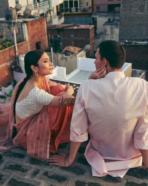 Romantic couple on a rooftop, woman in sari looks at man in a pink shirt with buildings in the background, bathed in warm sunlight.