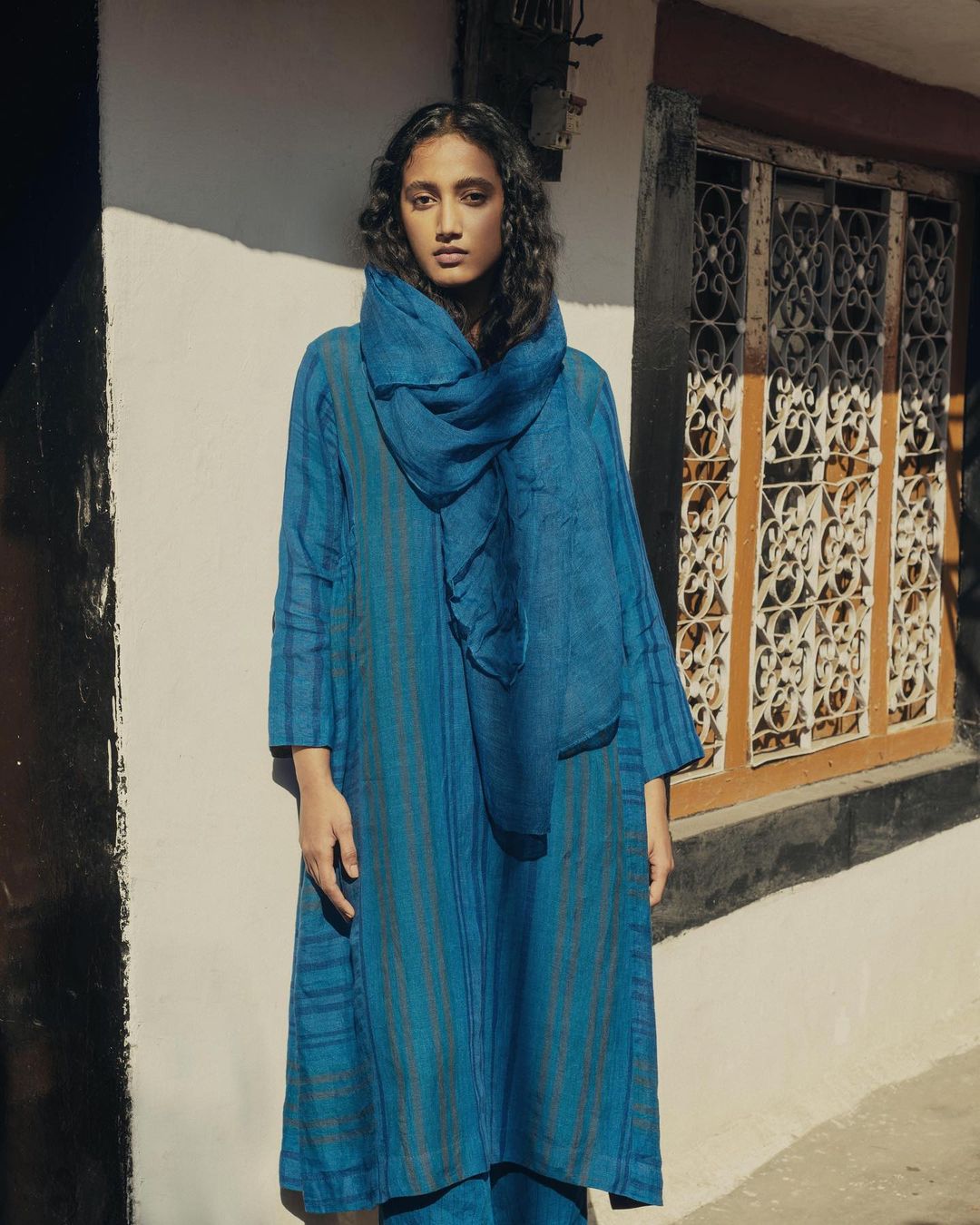 An East-Indian adult female stands outdoors next to a building wearing a long blue striped dress and scarf in eye-level medium shot.