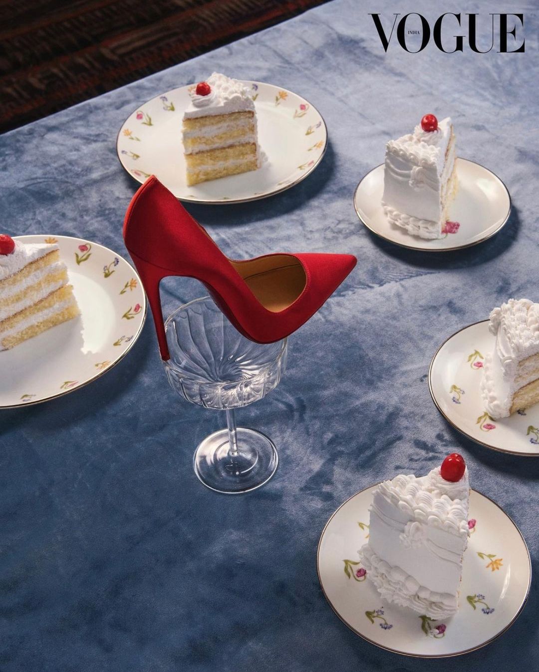 High-heeled shoe resting on a wine glass surrounded by cake slices, presented on a blue velvet surface, Vogue logo visible at top.