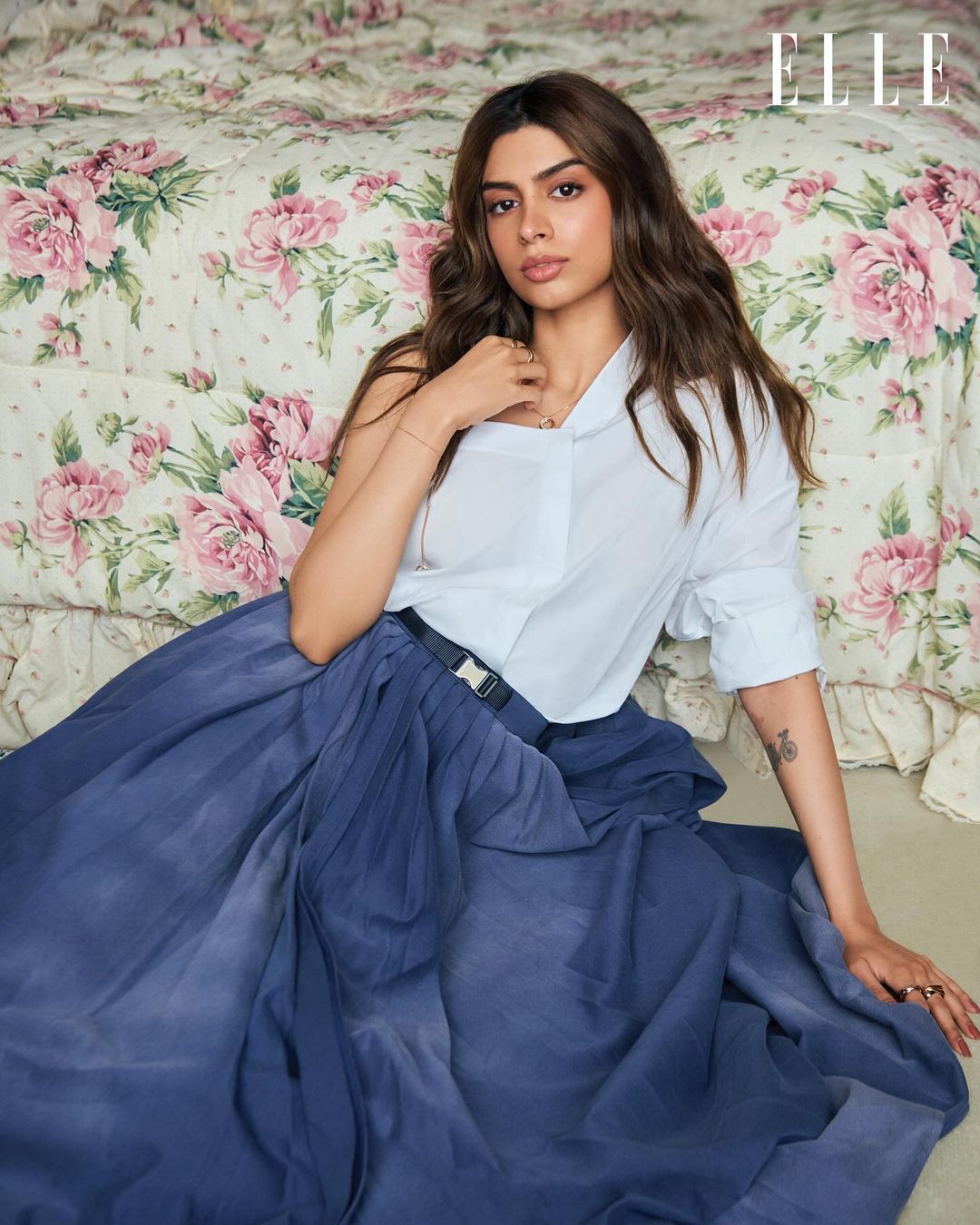 An adult female sits on a floral print bed with an oversized white dress shirt and a dark blue, flowing skirt with an Elle logo in the upper right.
