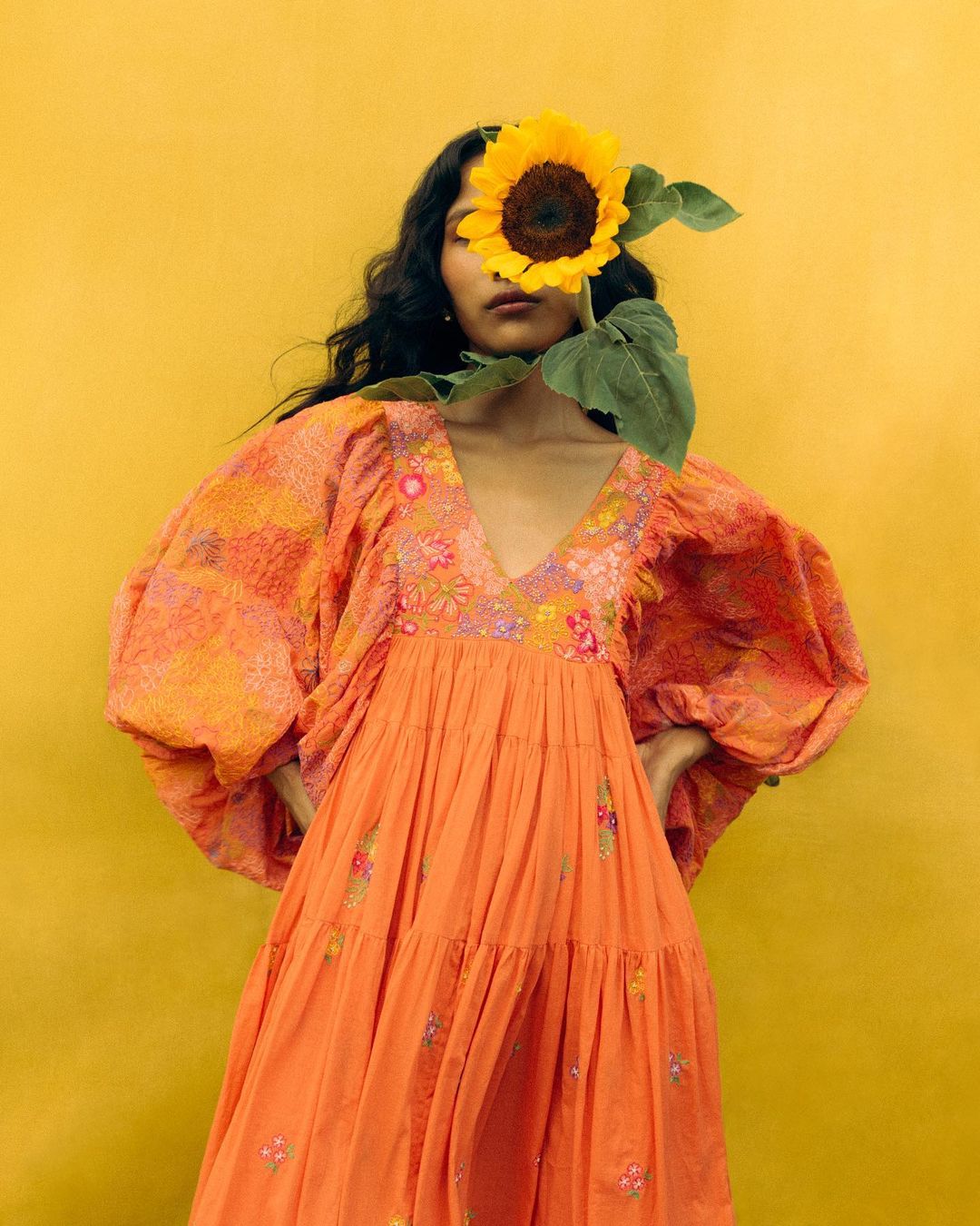 A woman in an orange dress poses with a sunflower covering her face against a yellow backdrop, creating a vibrant and artistic image.