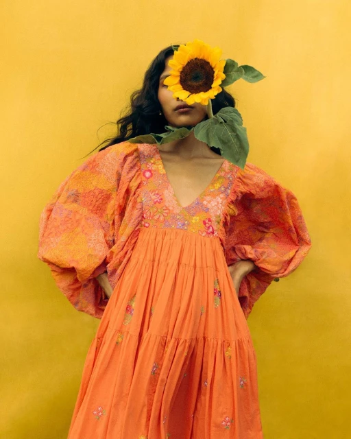 A woman in an orange dress poses with a sunflower covering her face against a yellow backdrop, creating a vibrant and artistic image.