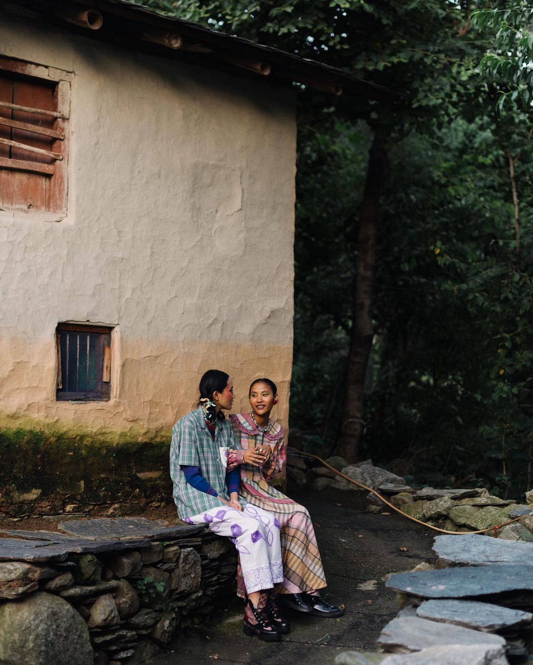Two women sit side-by-side on a stone wall near a building with trees in the background, dressed in patterned casual clothing.