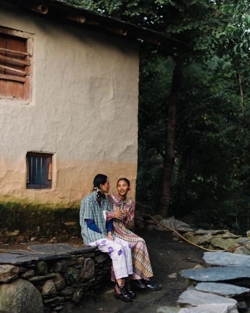 Two women sit side-by-side on a stone wall near a building with trees in the background, dressed in patterned casual clothing.