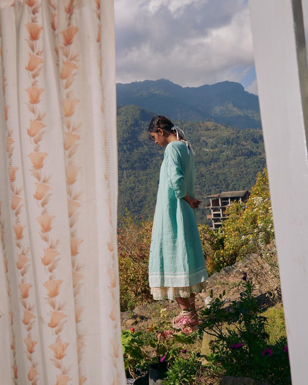 A young woman in a blue dress stands on a hilltop, framed by a window with floral curtains, overlooking a valley with mountains and sky.