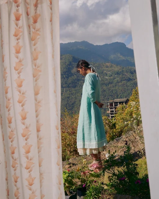 A young woman in a blue dress stands on a hilltop, framed by a window with floral curtains, overlooking a valley with mountains and sky.