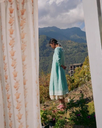 A young woman in a blue dress stands on a hilltop, framed by a window with floral curtains, overlooking a valley with mountains and sky.