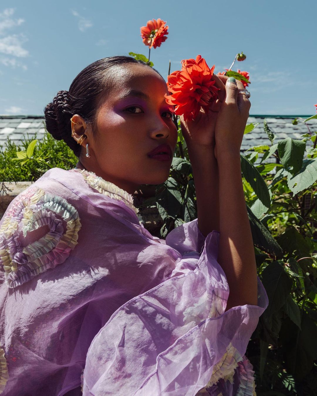 Portrait of woman with purple eyeshadow and lavender top holding a coral dahlia flower against a blue sky and green foliage background.