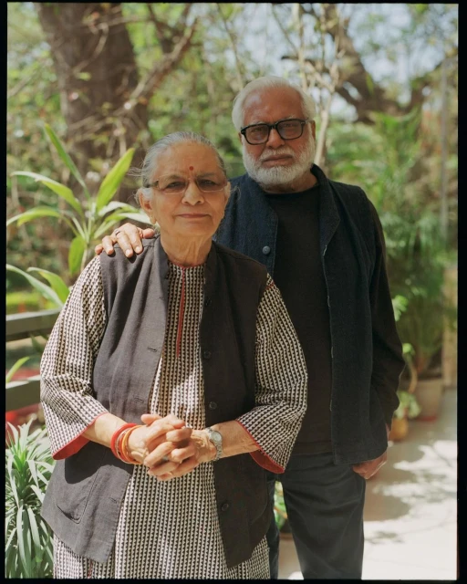 Older adult couple outdoors, posing in front of green foliage. The woman wears a patterned top with a black vest. The man has a beard.