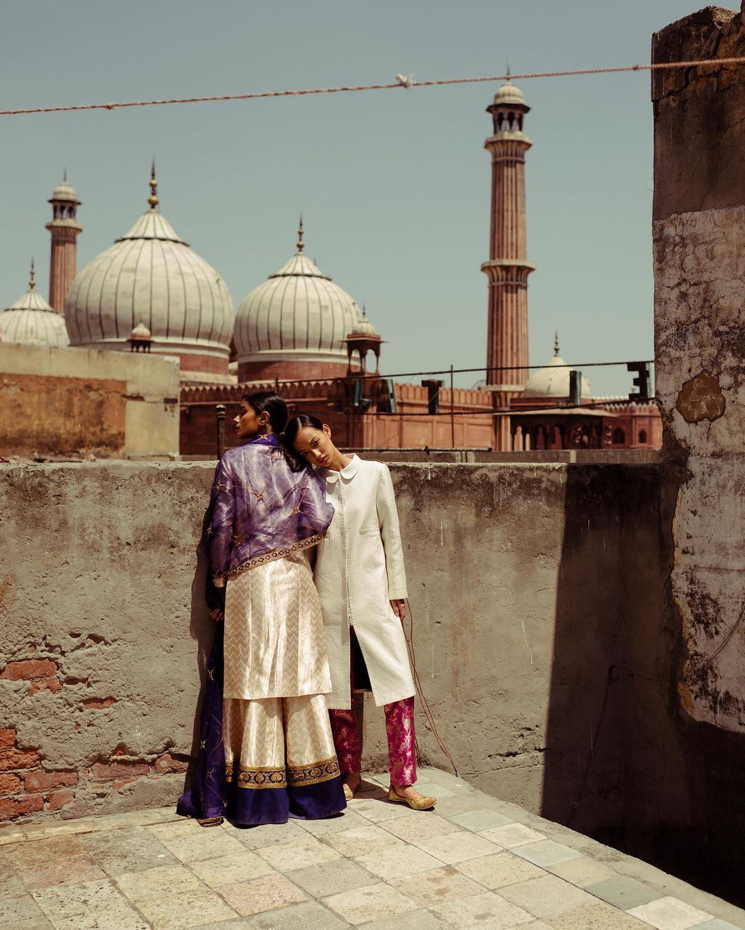 Two fashion models pose against a weathered wall, with a mosque in the background, wearing purple and white embroidered traditional Indian clothing.
