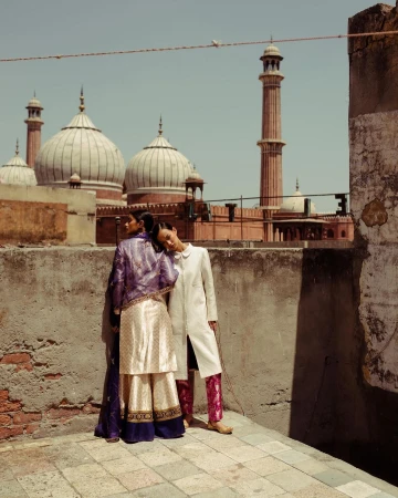 Two fashion models pose against a weathered wall, with a mosque in the background, wearing purple and white embroidered traditional Indian clothing.