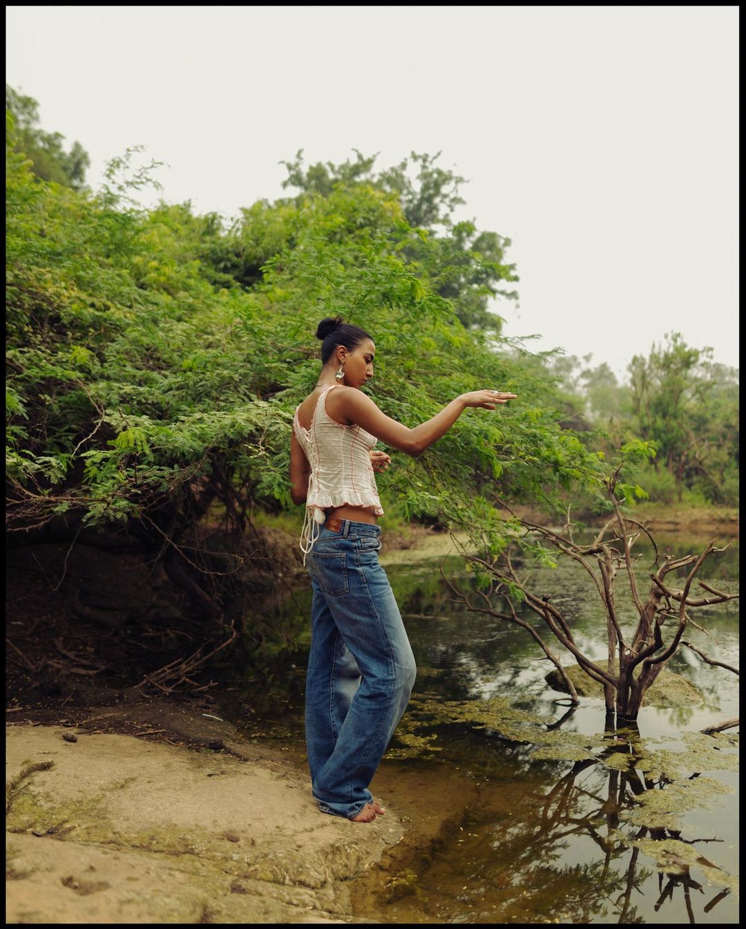 Woman in jeans and top standing barefoot near a pond surrounded by lush greenery and trees on an overcast day.
