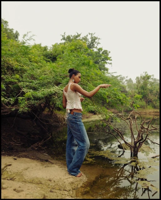 Woman in jeans and top standing barefoot near a pond surrounded by lush greenery and trees on an overcast day.