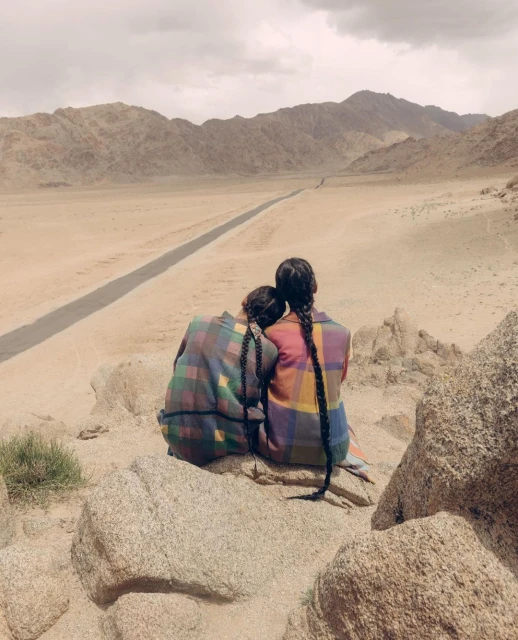 Two women with long braids wearing plaid tops sit on rocks overlooking a desert landscape and distant mountains on a cloudy day.