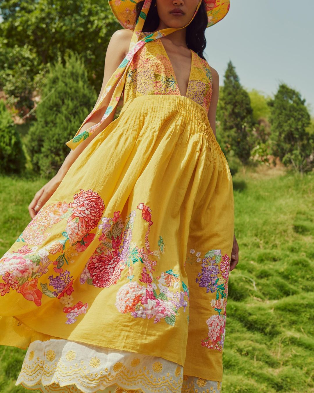 Woman in yellow floral halter dress and sun hat poses in a meadow with trees in the background on a sunny day, fashion shot.
