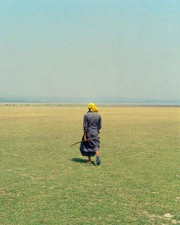 Person in headscarf walks through a grassy field toward a distant shore, with a hazy sky, carrying a long stick or branch.
