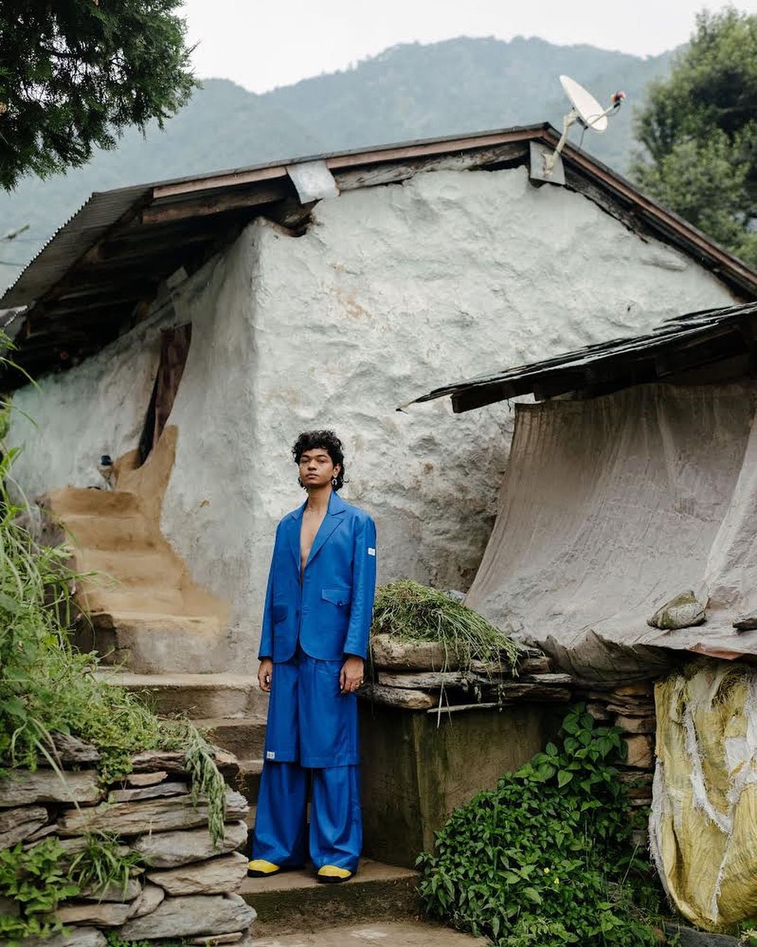 Portrait of person in bright blue suit and palazzo pants standing in rural village setting, traditional plastered house and lush greenery.