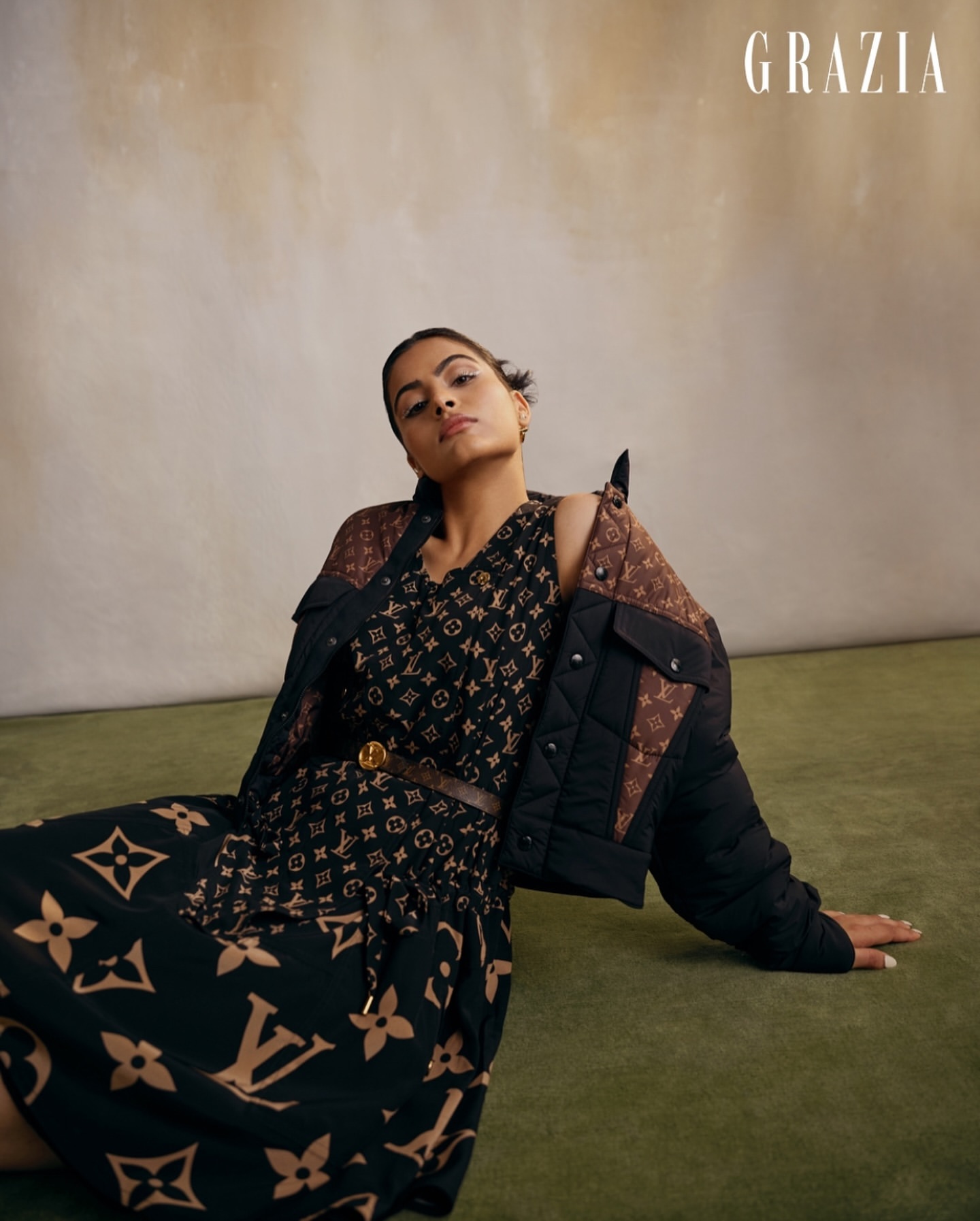Fashion editorial shot of a woman in a patterned dress and jacket, lounging on a green surface. Grazia magazine logo visible.