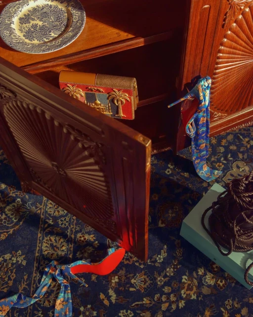 An intricately carved wooden cabinet displays a patterned plate and a decorative purse with a red heel and patterned scarf on a floral rug.