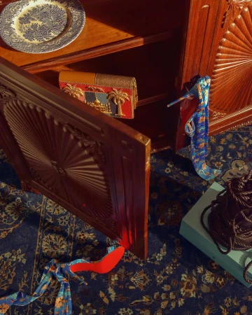 An intricately carved wooden cabinet displays a patterned plate and a decorative purse with a red heel and patterned scarf on a floral rug.