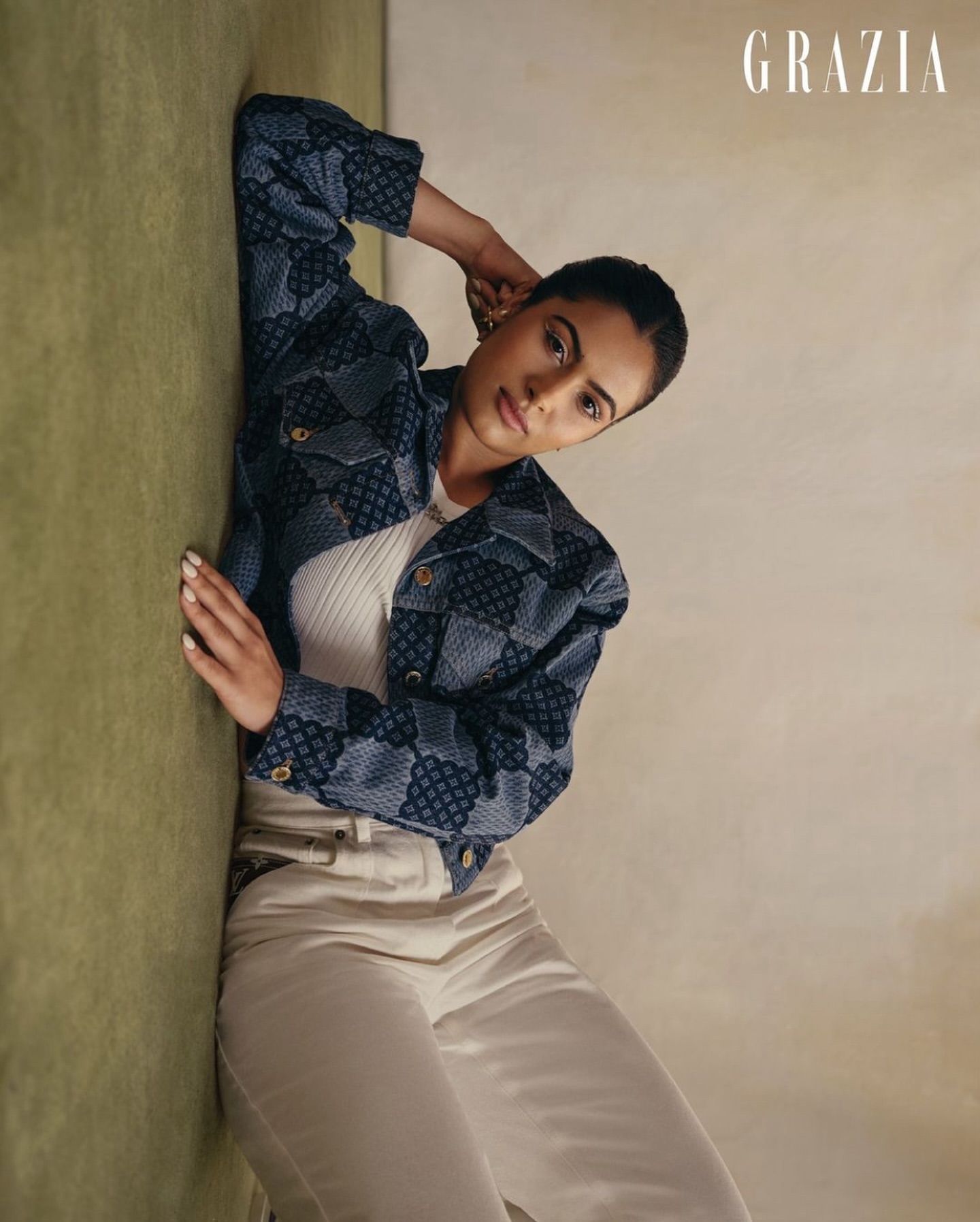 Attractive brunette woman poses in a patterned jacket and white pants, indoors with soft lighting in a vertical studio shot.