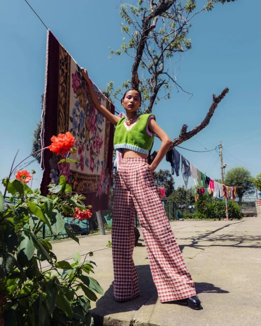 Woman in green top and checkered pants poses with rug outdoors on sunny day, framed by tree and clothesline in background.