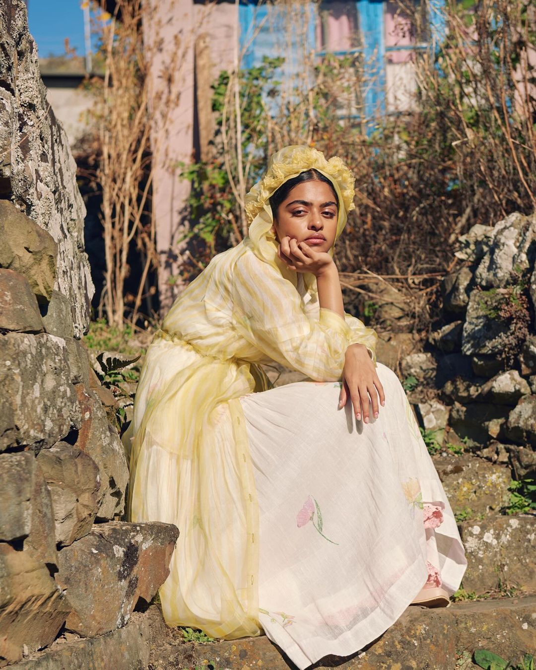 Woman in a yellow bonnet and patterned dress sits on stone steps in a rural setting, posing with hand to face in natural sunlight.