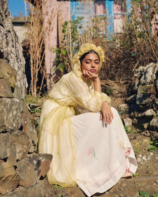 Woman in a yellow bonnet and patterned dress sits on stone steps in a rural setting, posing with hand to face in natural sunlight.