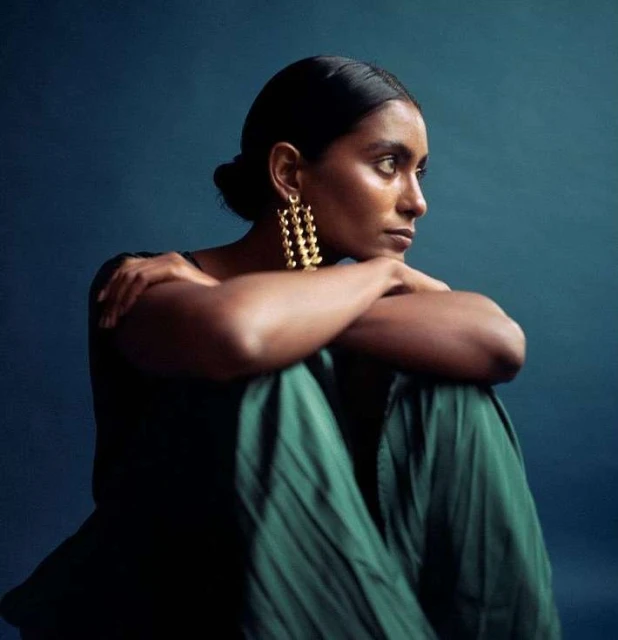 Portrait of a young adult woman with dark hair, wearing gold statement earrings and a green dress, against a dark teal backdrop.