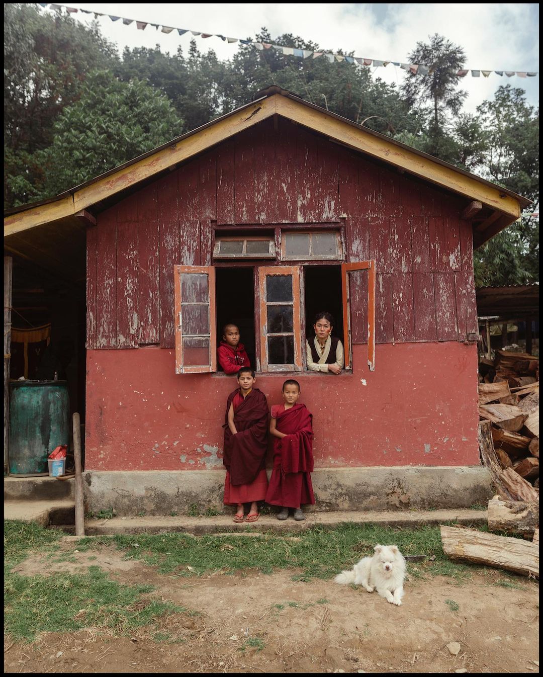 Group of children and an adult at the window of a red house with a white dog lying on the ground in a rural environment.
