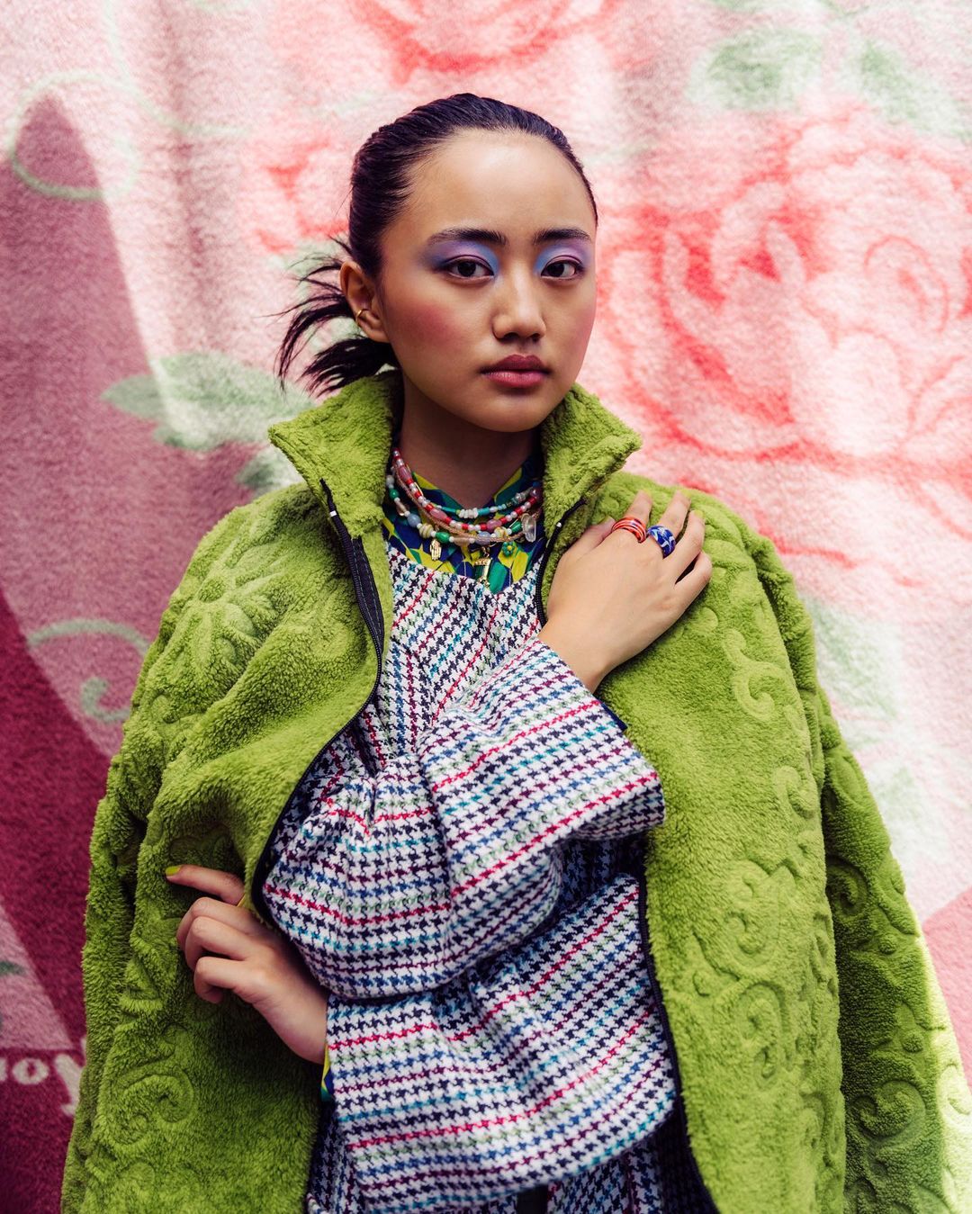 Portrait of an Asian woman with colorful makeup and jewelry, wearing a green coat against a pink floral patterned background.