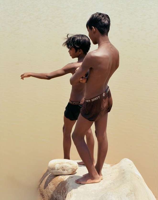 Two bare-chested boys stand on a rock in a river, one with arms outstretched, the other looking on, captured in a medium outdoor shot.